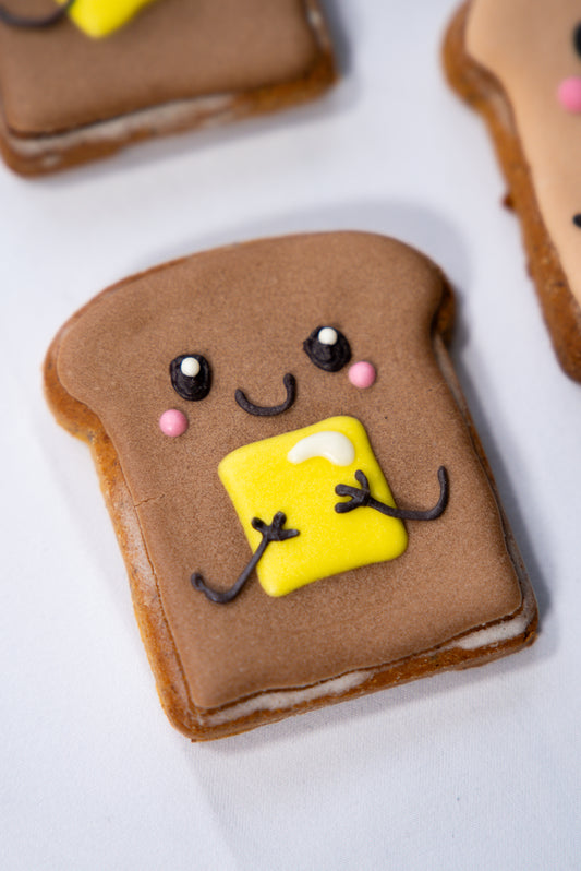 Cookie shaped like a piece of toast with a smiley face and a square of yellow fondant on top.